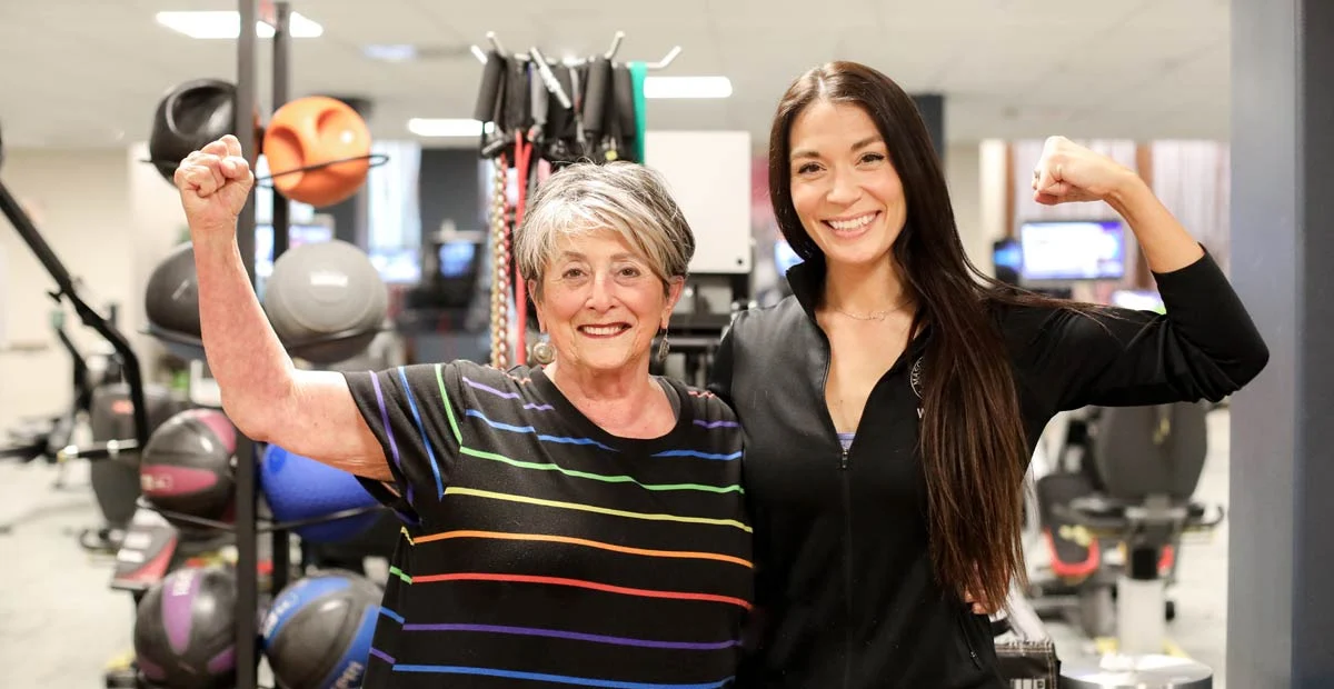 Two women, a resident and wellness center staff member, standing together and smiling at the camera while showing off arm muscles.