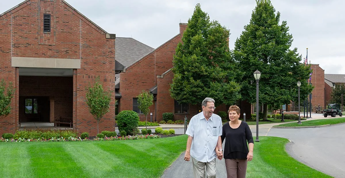 Senior couple holding hands while walking outdoors together. Large brick buildings are in the background behind them.