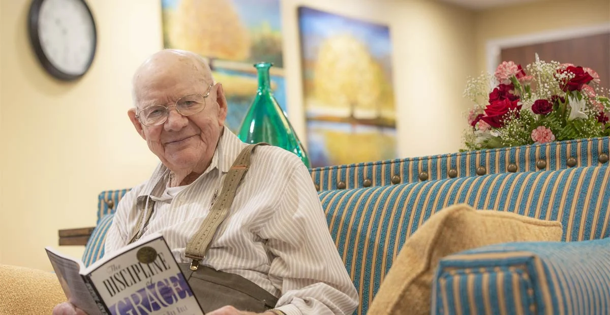 Senior man smiling with a book open in his hands while seated on a colorful couch.