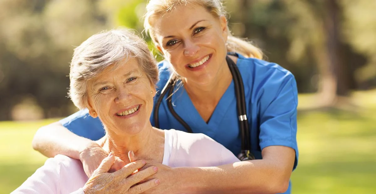 Senior woman being hugged by a smiling nurse outdoors.
