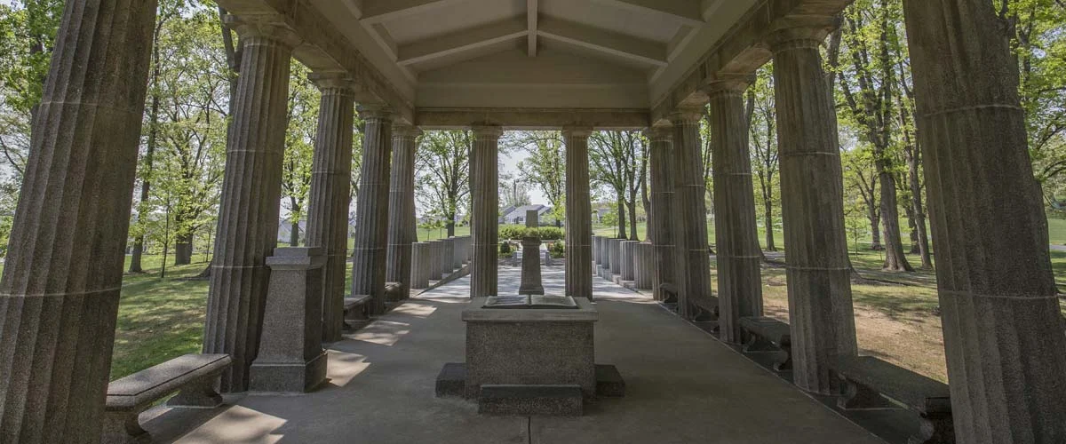 Hero_Veterans-Grove-04 View from inside Veteran's Grove loggia with path leading outside and trees surrounding the area.
