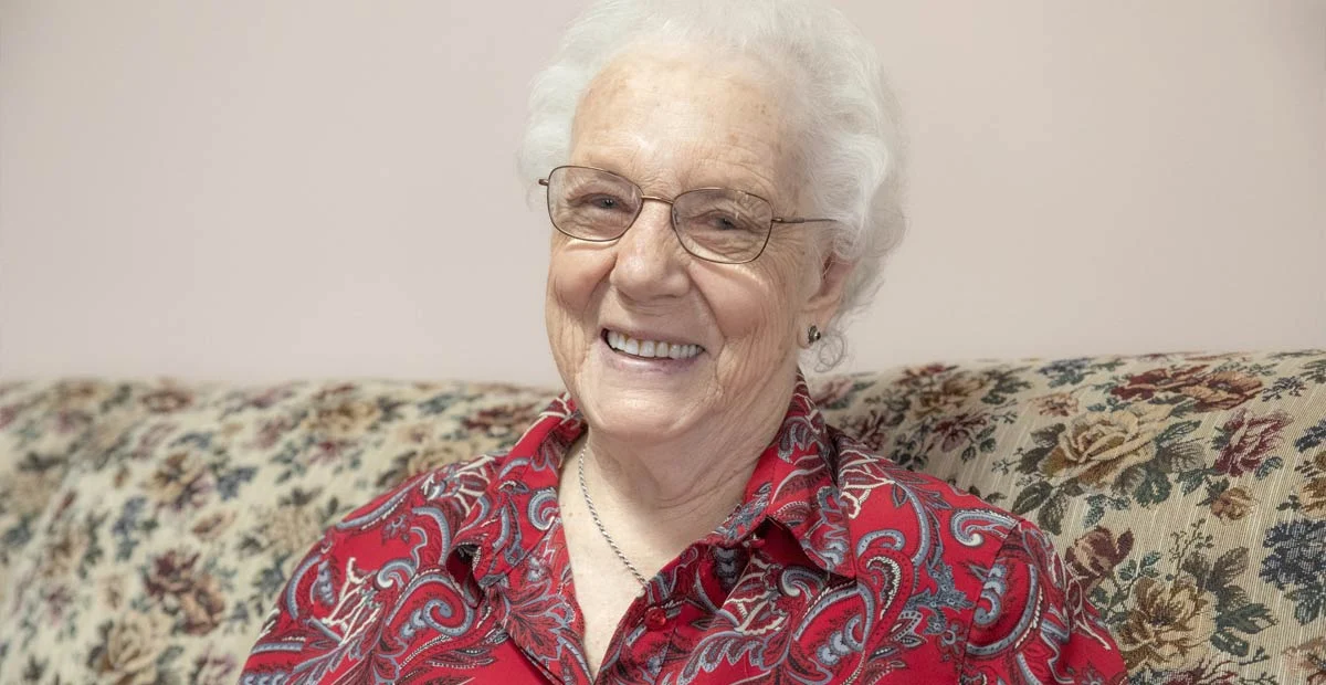 Mrs. Kolmus, a smiling senior woman in glasses seated on a couch.