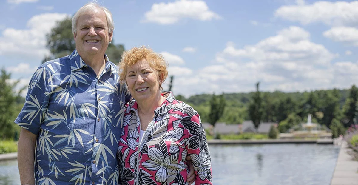 Smiling senior couple in floral shirts standing outdoors in a large garden.