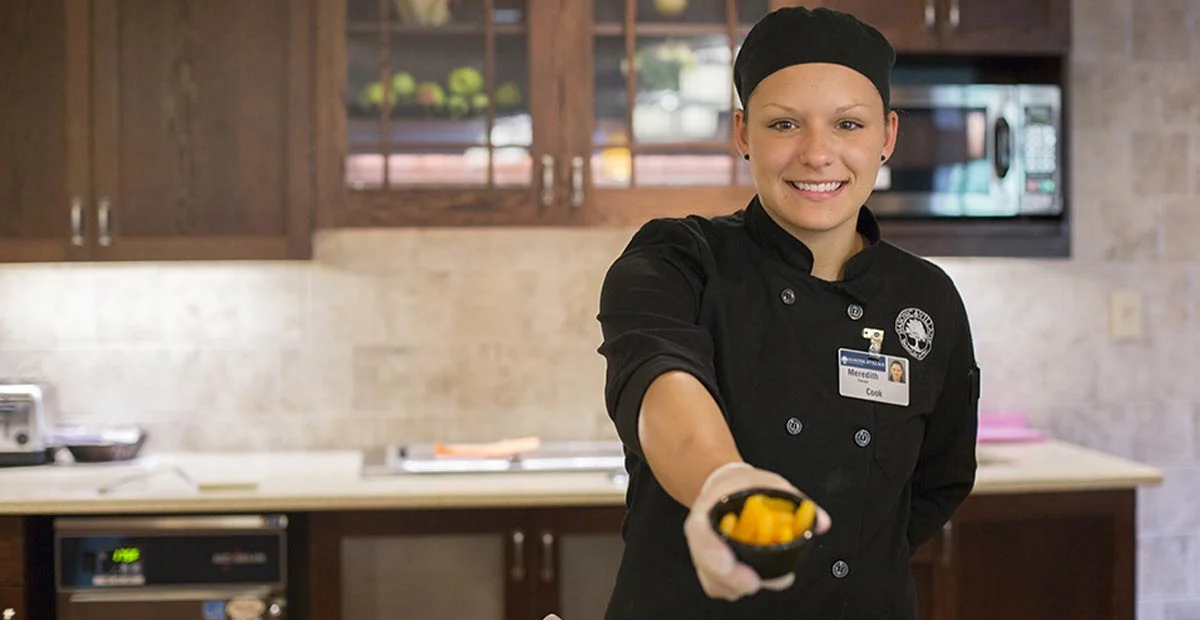 Smiling young woman in chef's uniform holding out sliced fruit in a cup.