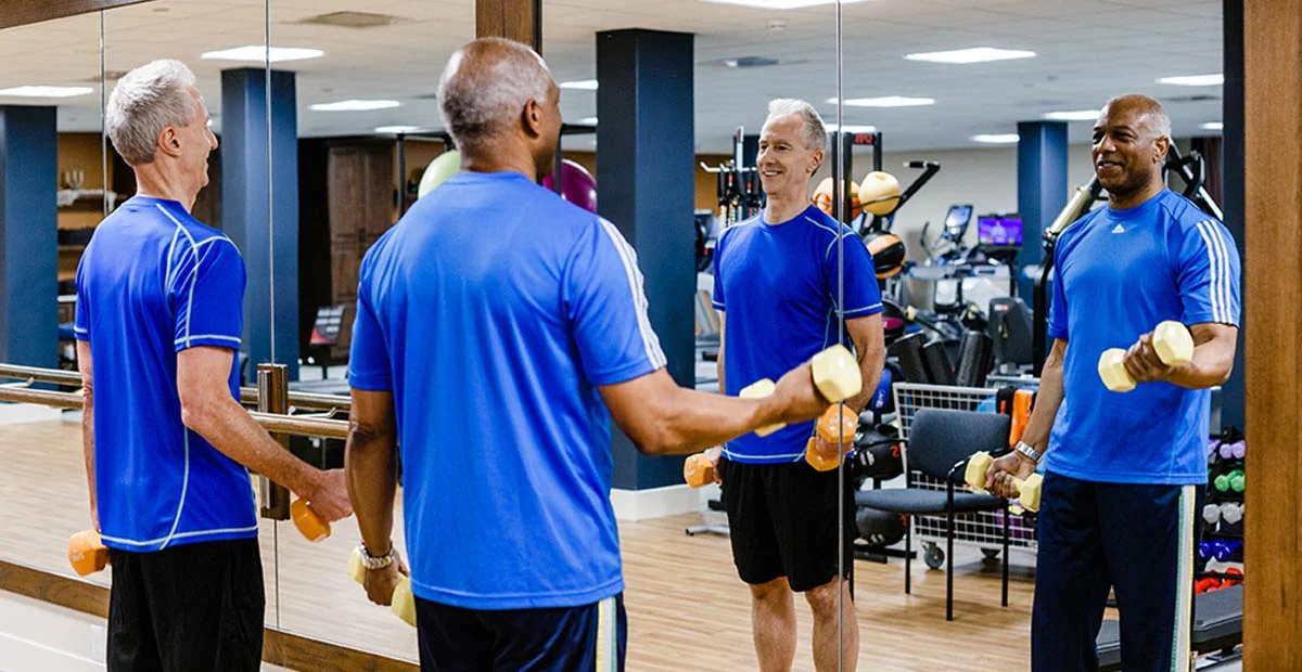 Smiling senior men lifting weights together while looking in a mirror.
