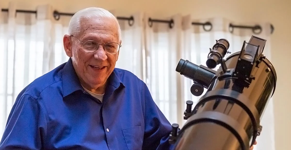A smiling senior man in glasses standing next to a large, black telescope.