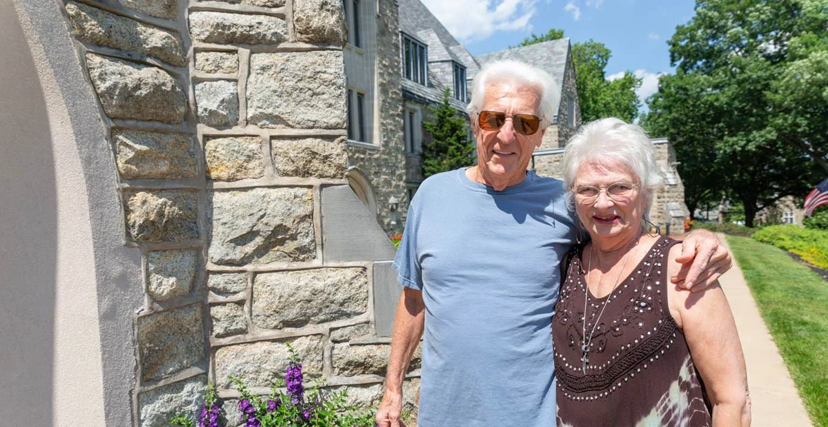 Don and Pat Gibble, a smiling senior couple standing outside of large, stone building.