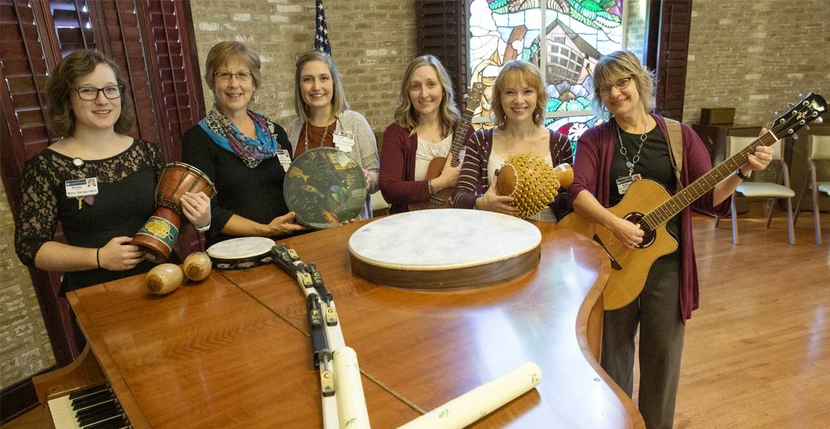 Smiling group of music therapists with their instruments while gathered around a piano.