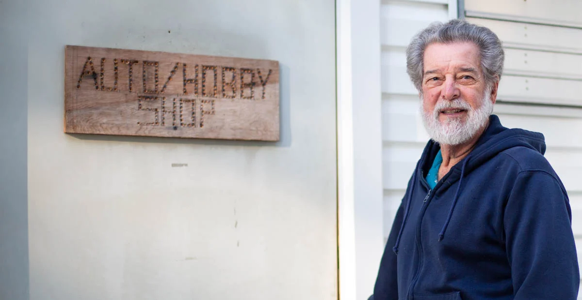 Smiling senior man standing by wood sign on a white building, reading "Auto/Hobby Shop."