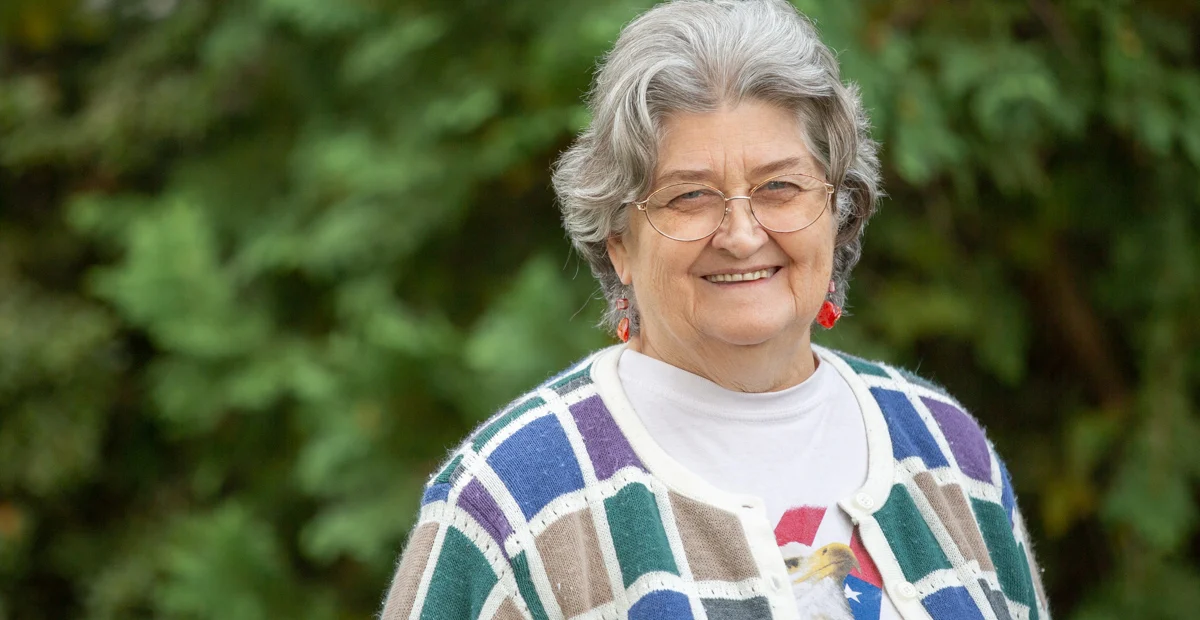 Evelyn Pollock, a smiling senior woman with glasses and short hair wearing a colorful cardigan. She's standing outdoors with greenery behind her.
