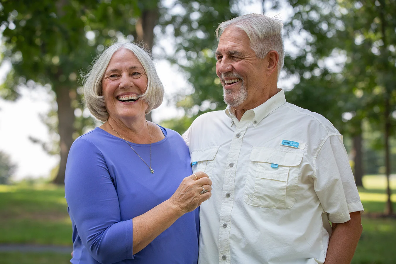 Smiling senior couple standing together outdoors. Sunlit trees are in the background behind them.