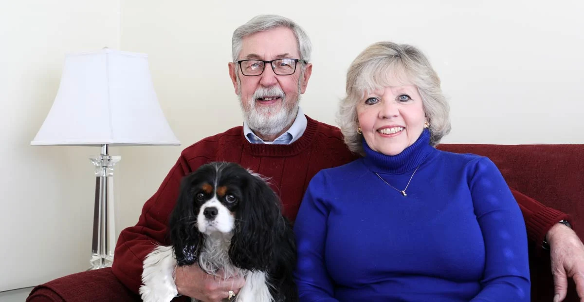 Smiling senior couple seated on a red couch with their King Charles Spaniel.