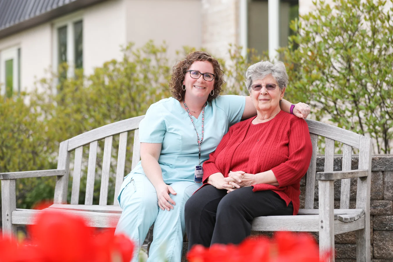 Smiling senior woman sitting with personal care aide on a bench outdoors. The personal care aide has her arm around the senior woman's shoulders.