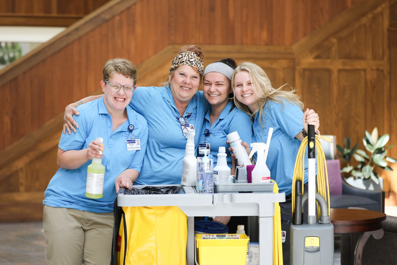 Smiling group of housekeeper staff members in blue with a cleaning cart.