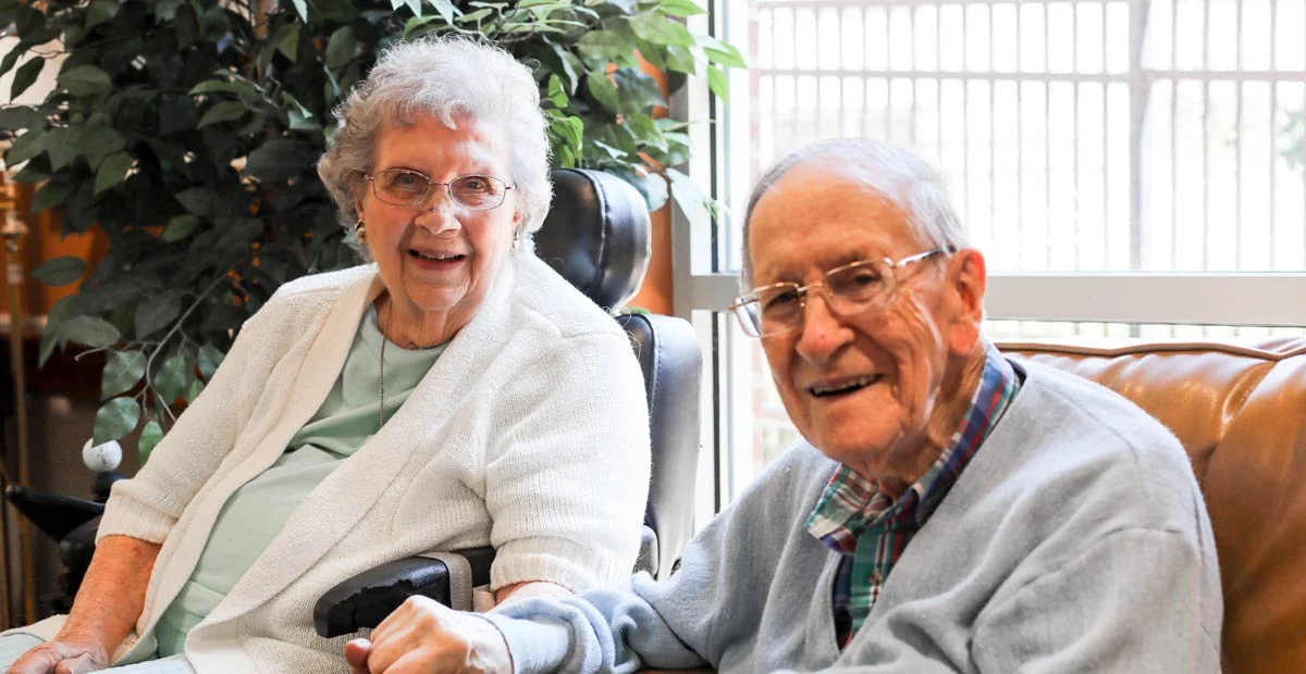 Mary and Jim Whitacre, a smiling senior couple holding hands while seated in their home.