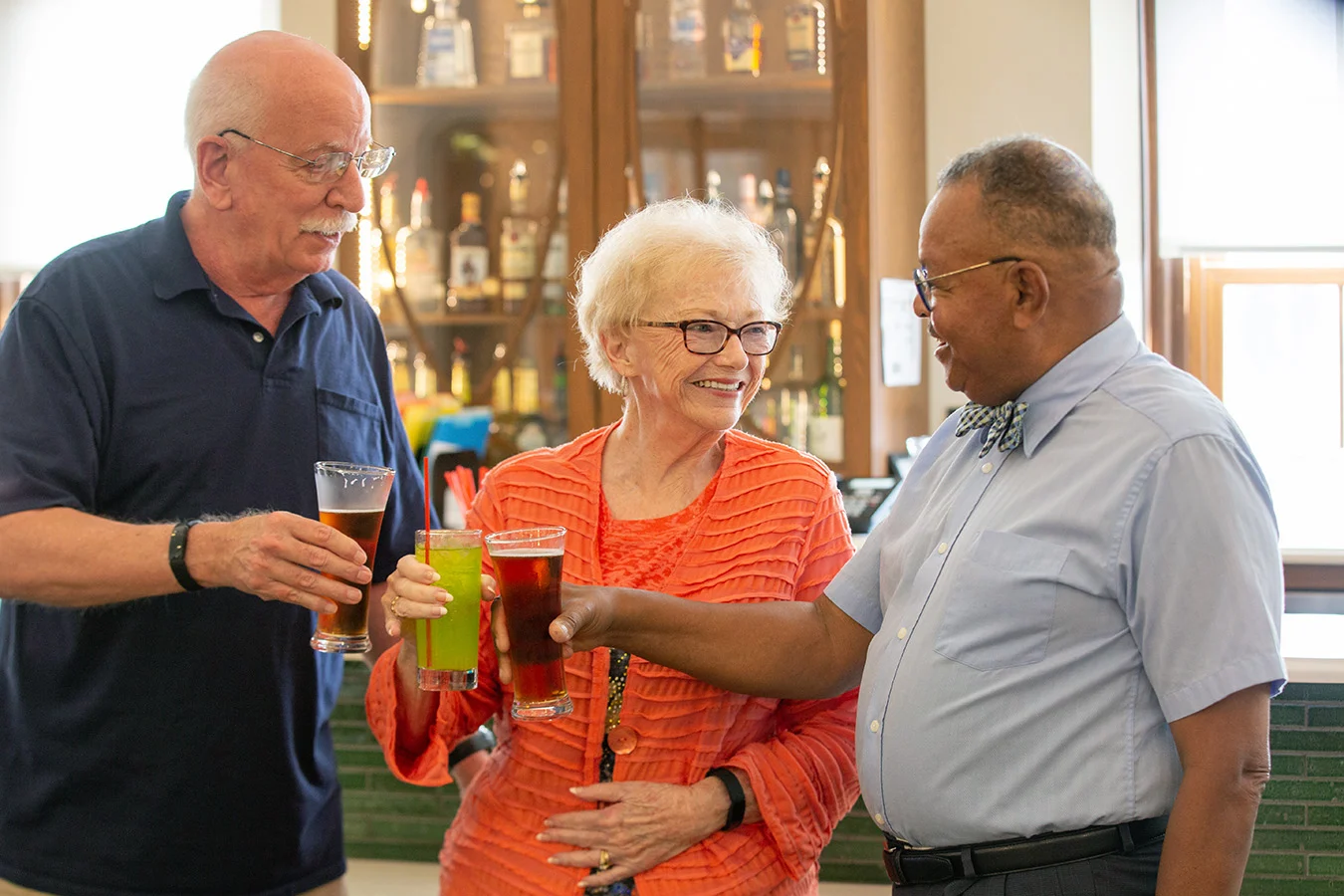 Three smiling seniors raising their drinks while standing in front of well-lit bar.