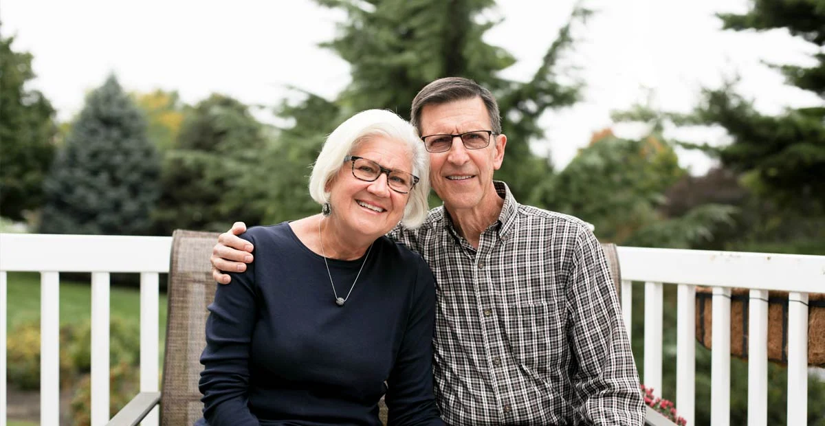 Brenda and Mike Evanko, a smiling couple with arms around each other seated on bench outdoors.