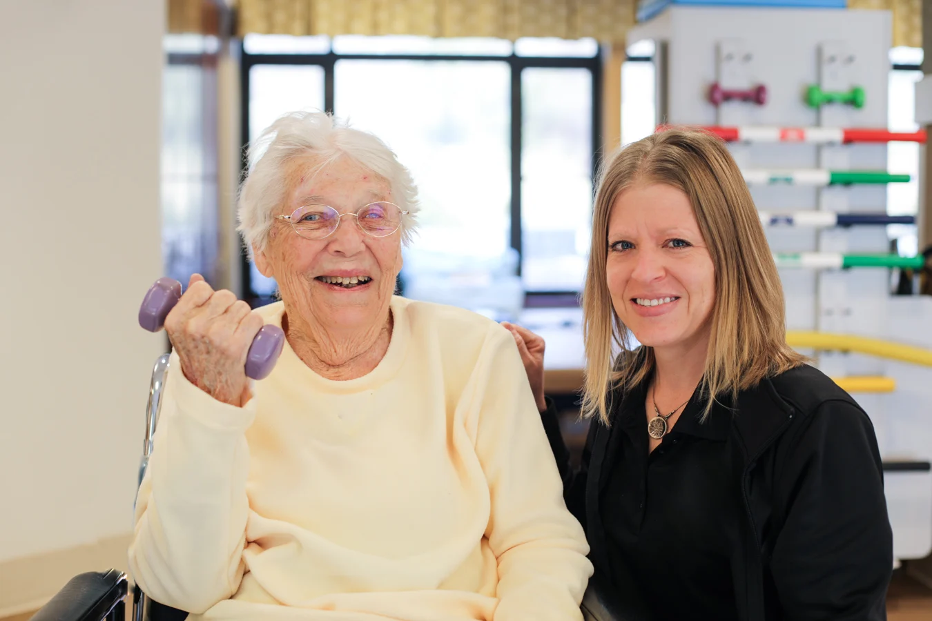 Physical Therapist seated next to smiling senior woman lifting a weight.