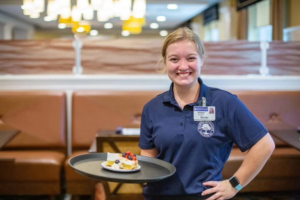 Smiling server holding a tray with a slice of cake in dining hall.