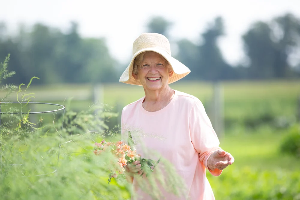 858A2353 Smiling senior woman in a large-brimmed hat holding flowers while outdoors in garden.