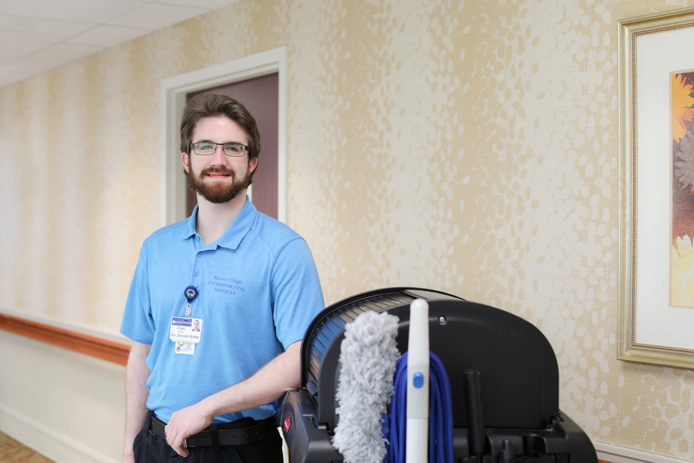 Smiling young man with glasses wearing a blue Environmental Services polo while standing in a hallway.