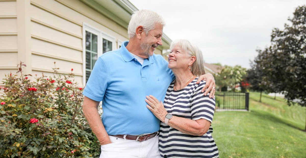 Gary and Debbie Morris, a smiling couple with arms around each other standing together in front of their home.