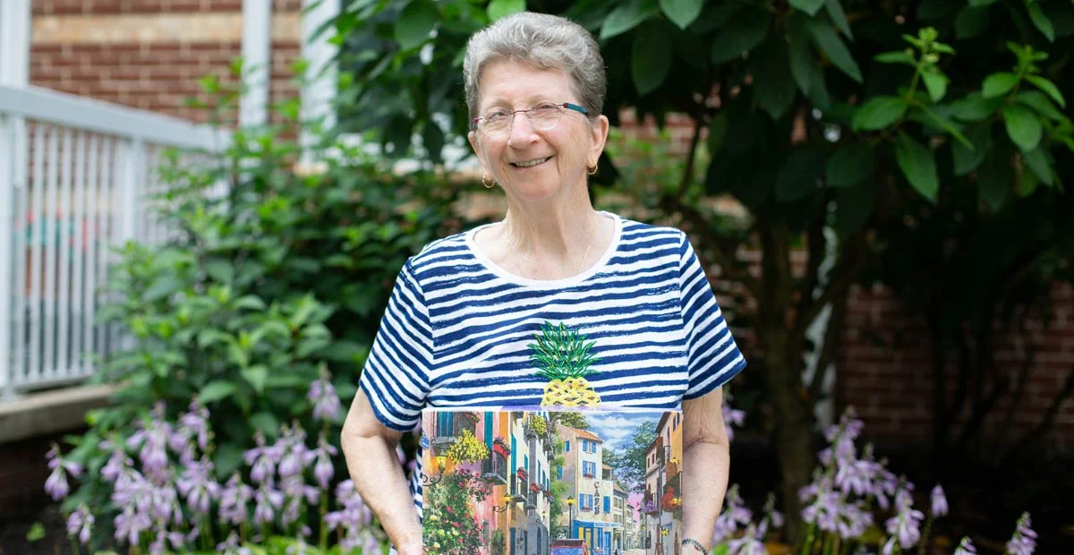 Karen Kostenbauder, a smiling senior woman with glasses holding up a painted canvas while standing outdoors.