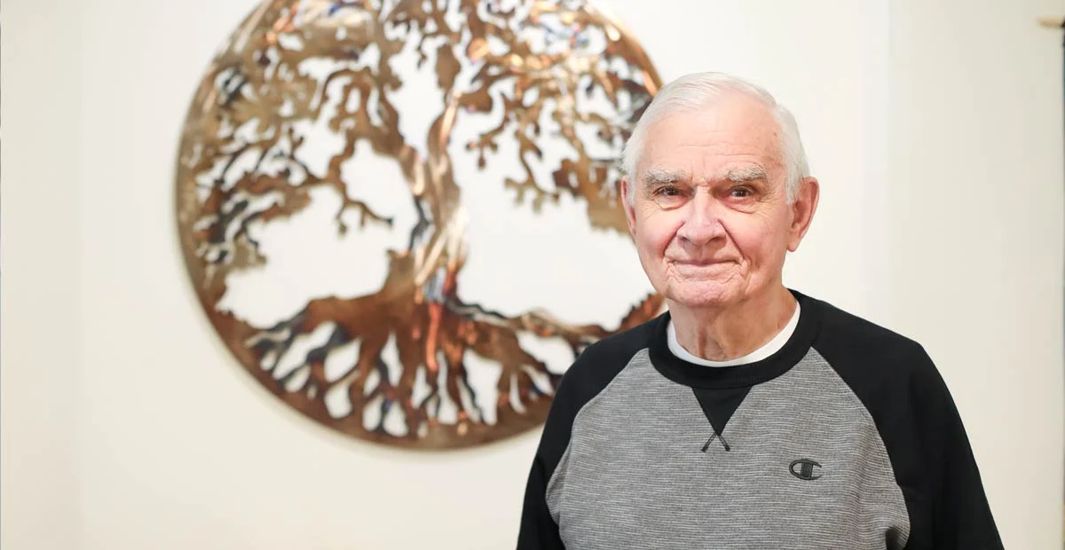 Chuck Armstrong, a smiling senior man standing in front of wall art cutout of a tree.