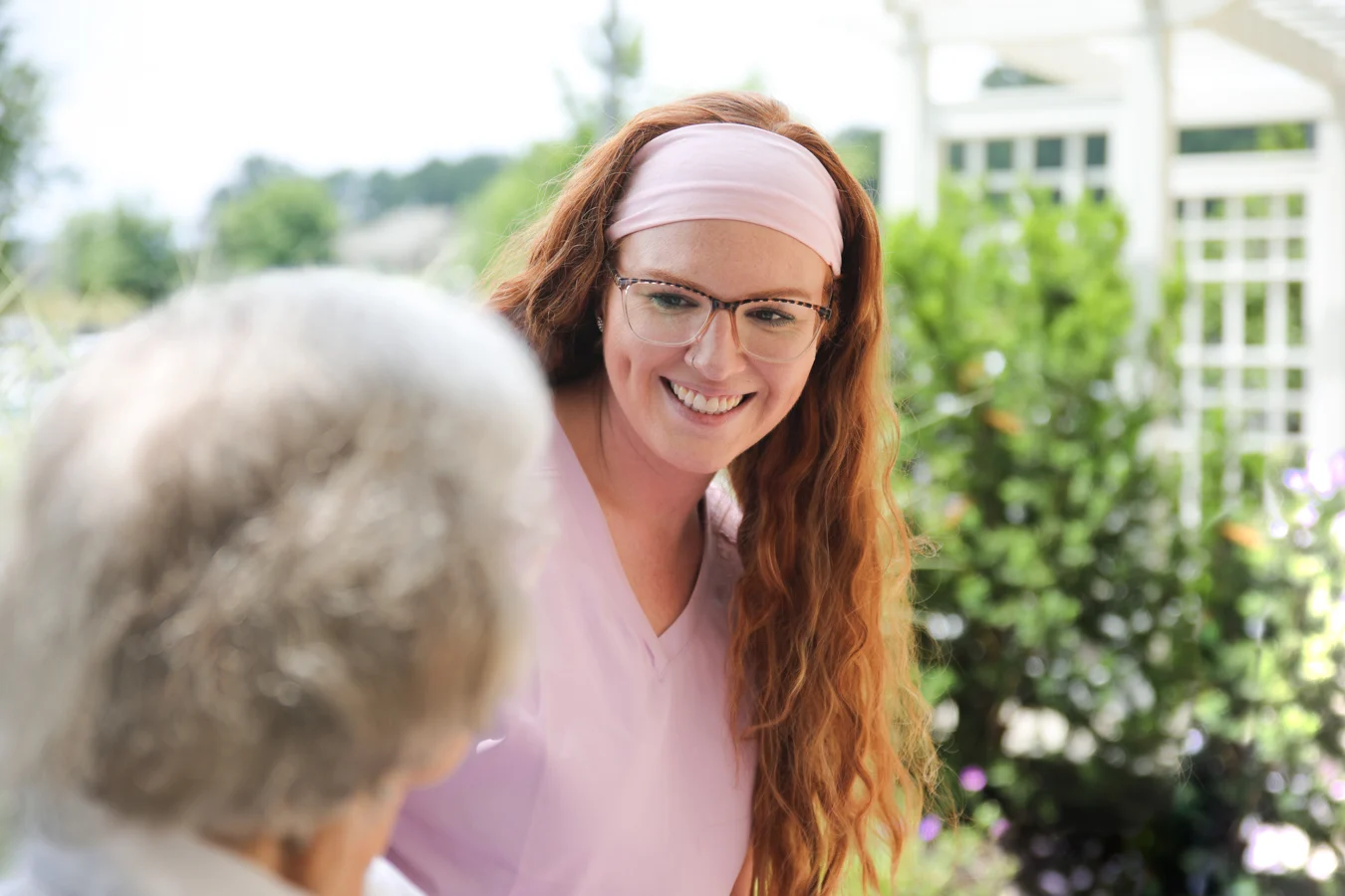 Nurse smiling at senior woman who is facing away from the camera while they are seated outdoors together.