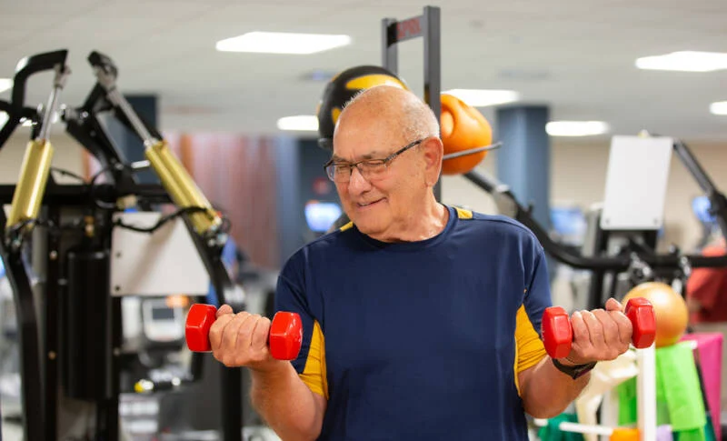 858A2305-web-800×485 Smiling senior man lifting weights inside of wellness center.