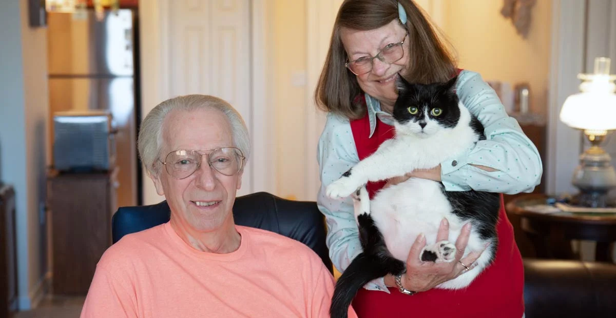 Robert and Bonnie Weikert, a smiling couple in their home. Bonnie is standing next to Robert holding the couple's cat.