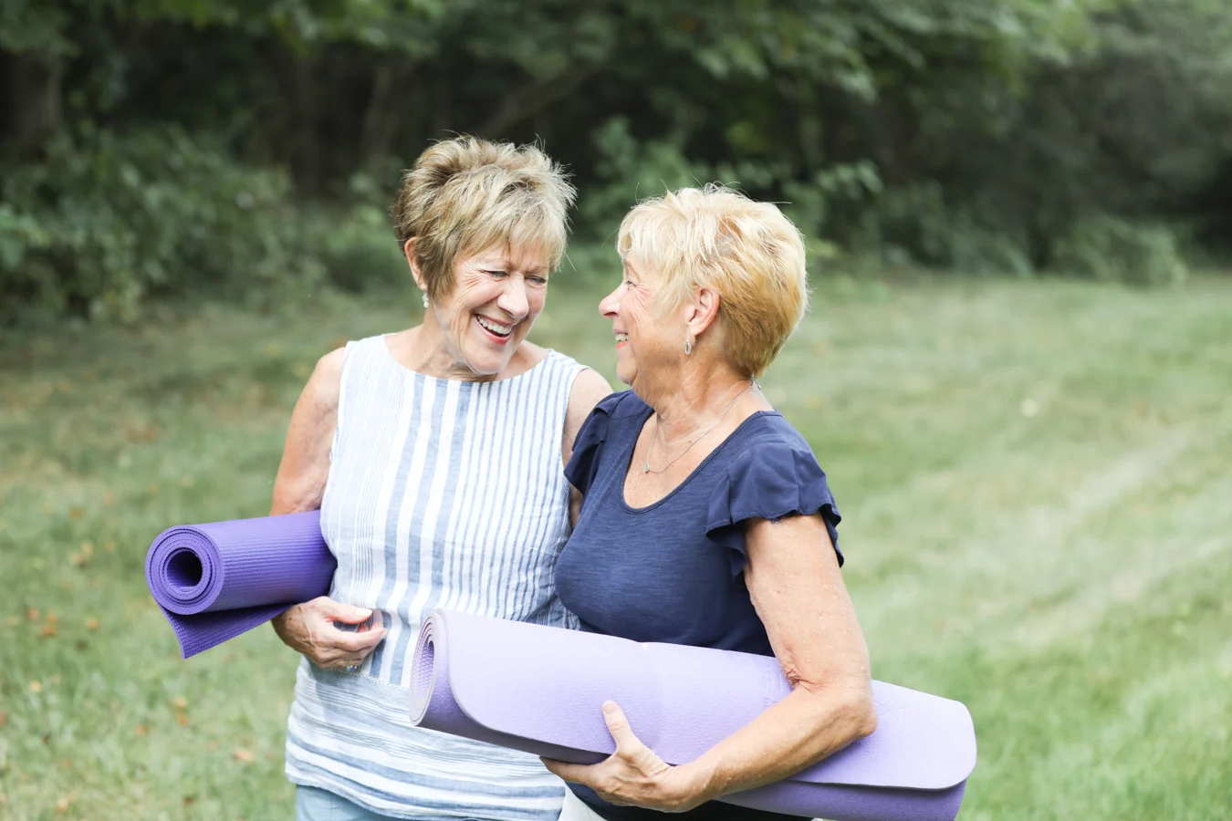 Two senior women smiling at each other and walking outdoors with their yoga mats.