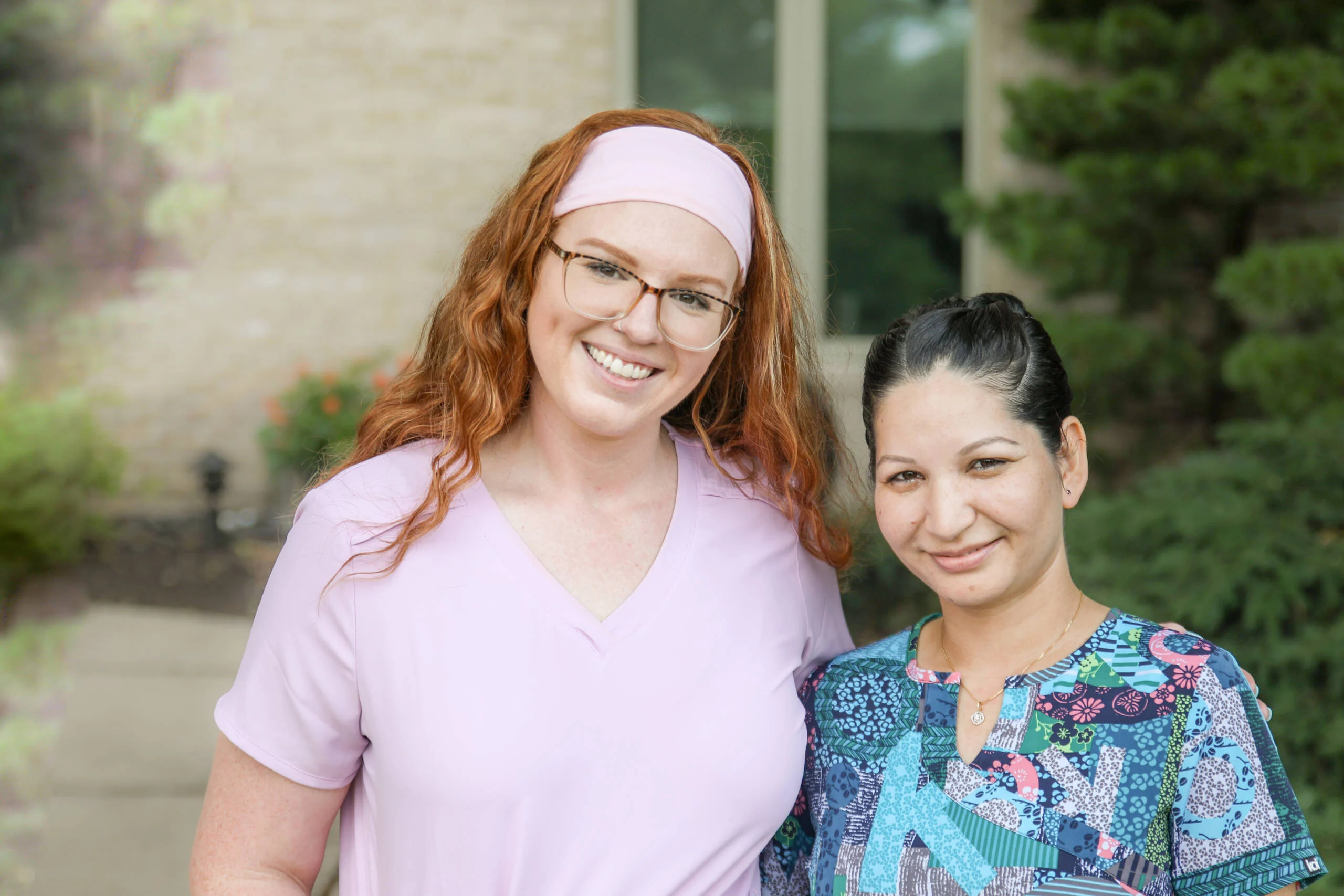 Two smiling young women dressed in scrubs while standing outdoors together.