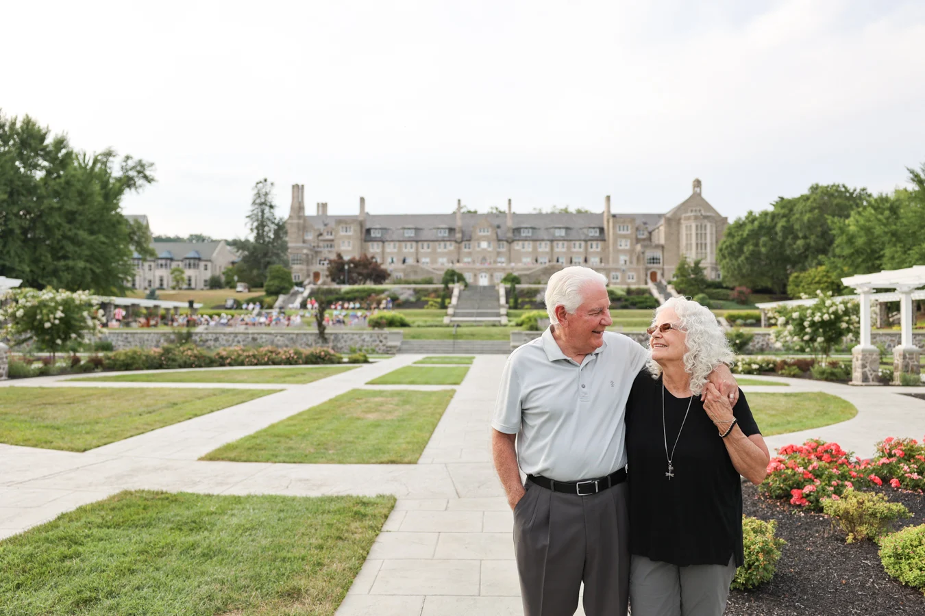 Smiling senior couple walking with arms around each other outdoors in garden, a large stone building is behind them in the distance.