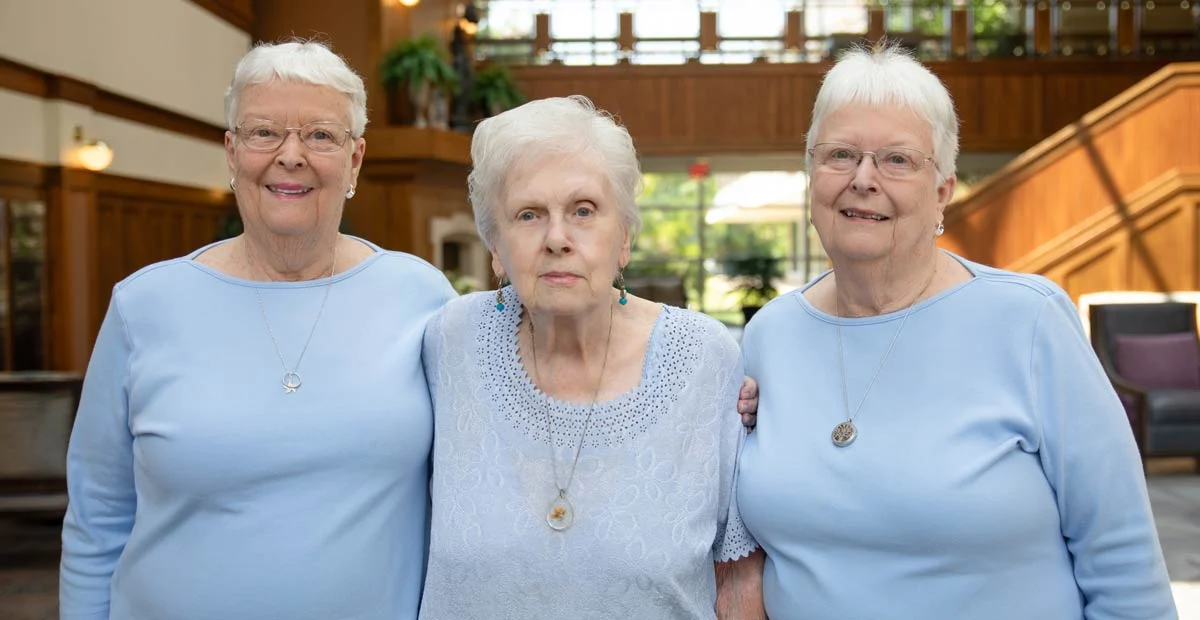 Group of three smiling senior women dressed in blue with arms around each other, standing indoors.