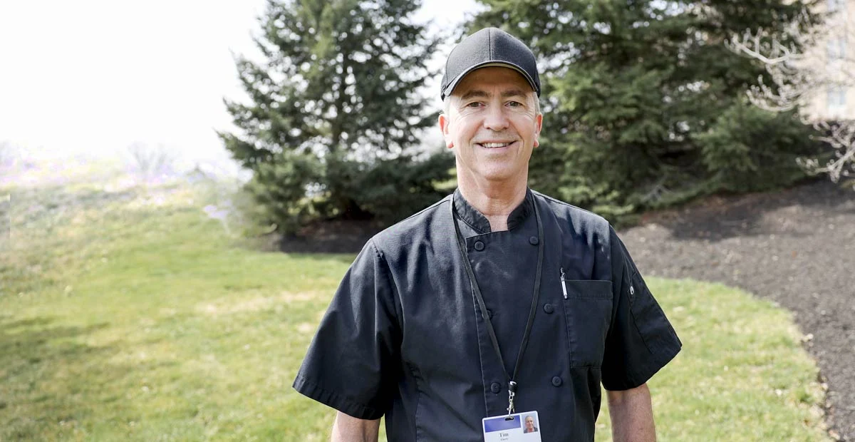Smiling man in black hat and chef's outfit standing outdoors.