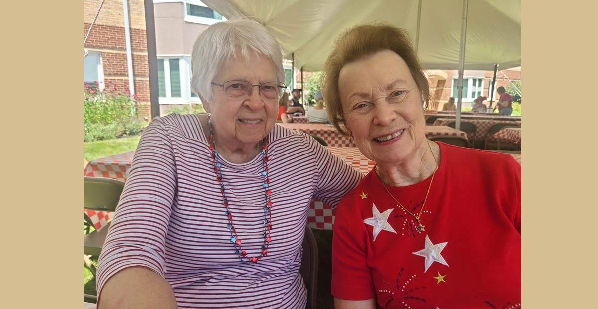 Nancy and Iola, two smiling senior women seated at a picnic table together under a tent.