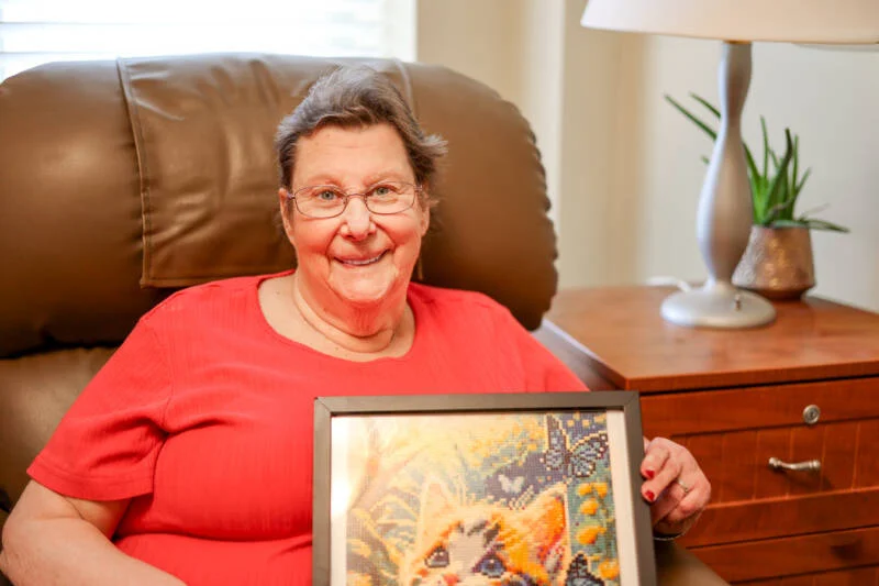 Smiling senior woman seated in a chair while holding up a piece of art.