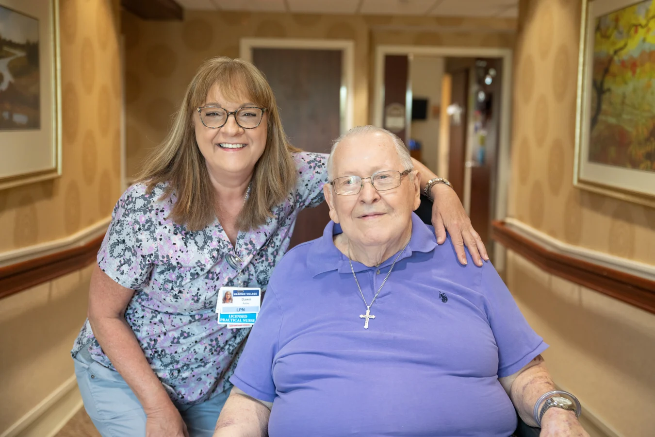 Smiling nurse standing with her arm around smiling senior man seated in a hallway.