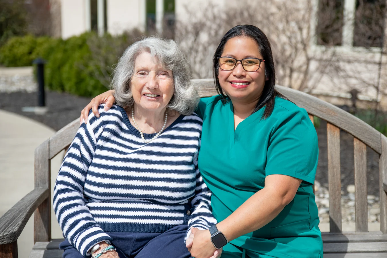 2V9A7324-Edit Smiling senior woman holding hands with a nurse while both are seated together on a bench outdoors.