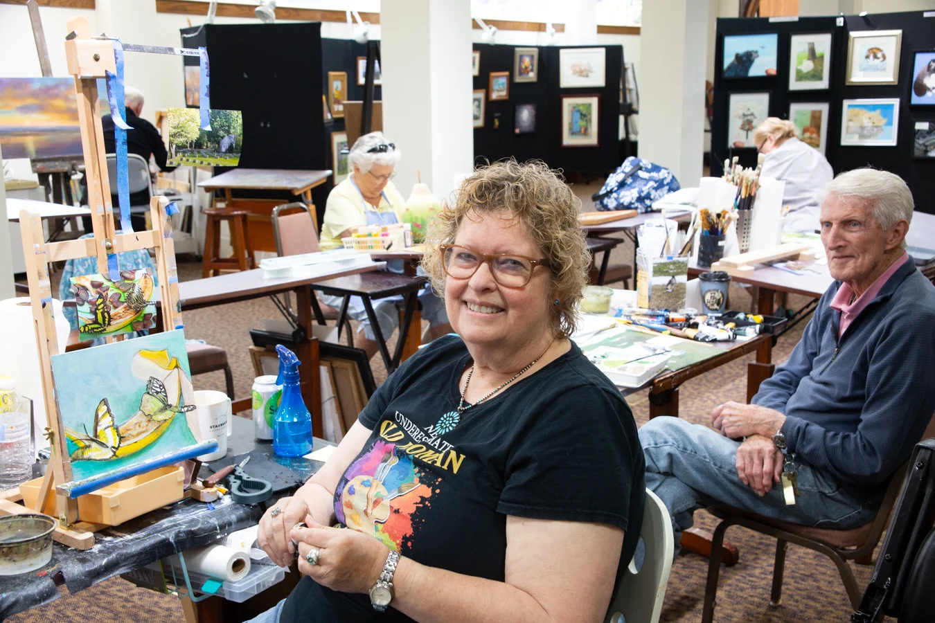 Smiling senior woman seated in art studio with her painting behind her. Others are working on their projects behind her.