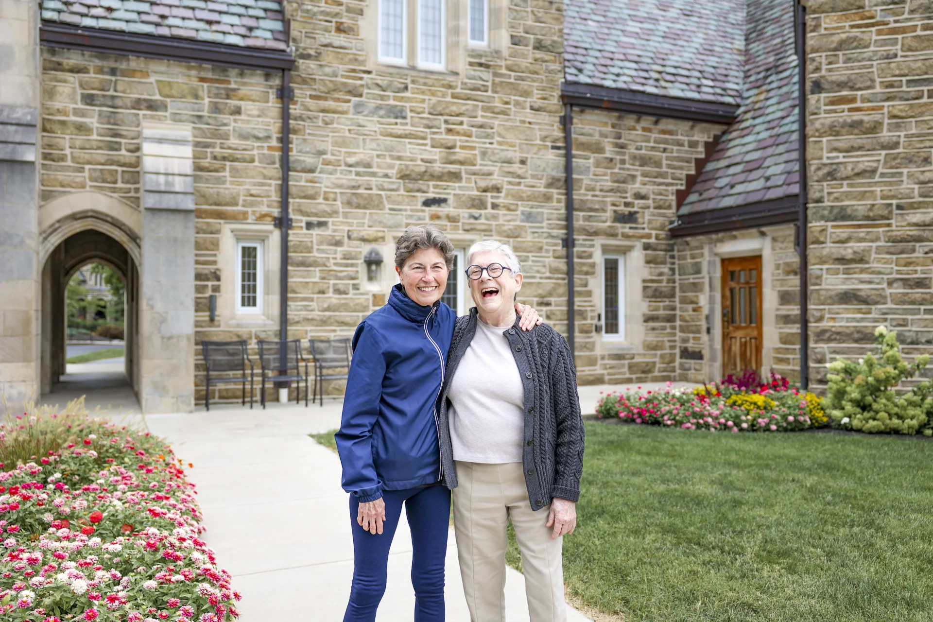 Two smiling senior women with arms around each other standing in front of a stone building outdoors.