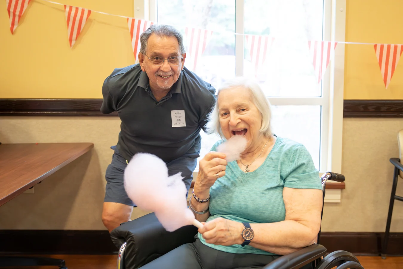 Smiling senior man and woman posed together indoors. The woman is seated in a wheelchair and eating a stick of cotton candy.