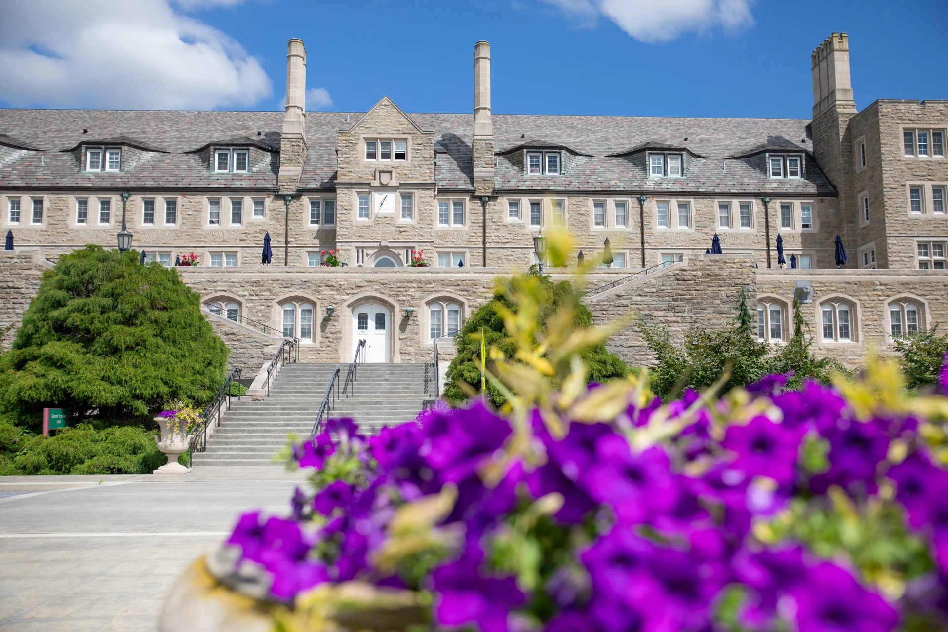 2V9A9194 Large, stone building with staircase leading up to it, framed by flowers and shrubs in the foreground.