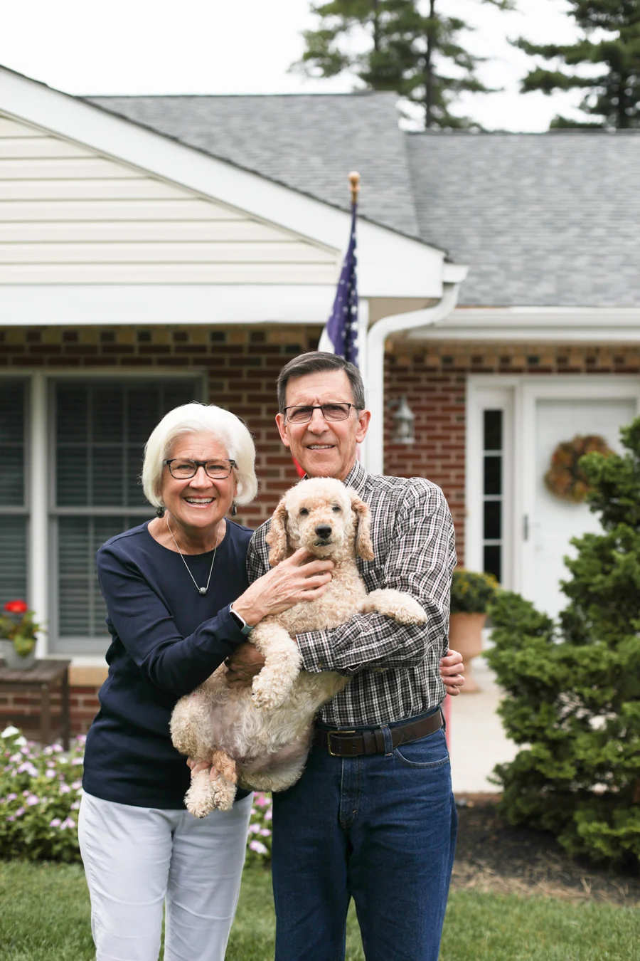 Smiling senior man and woman holding a dog while standing in front of their home.