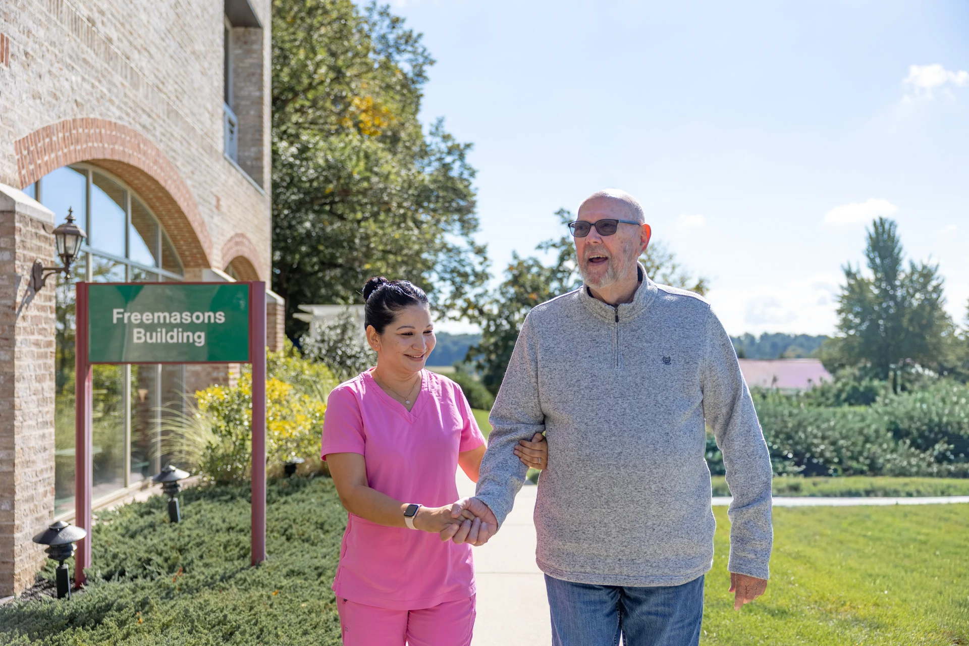 1W8A0917 Smiling senior man walking with personal care aide outdoors.