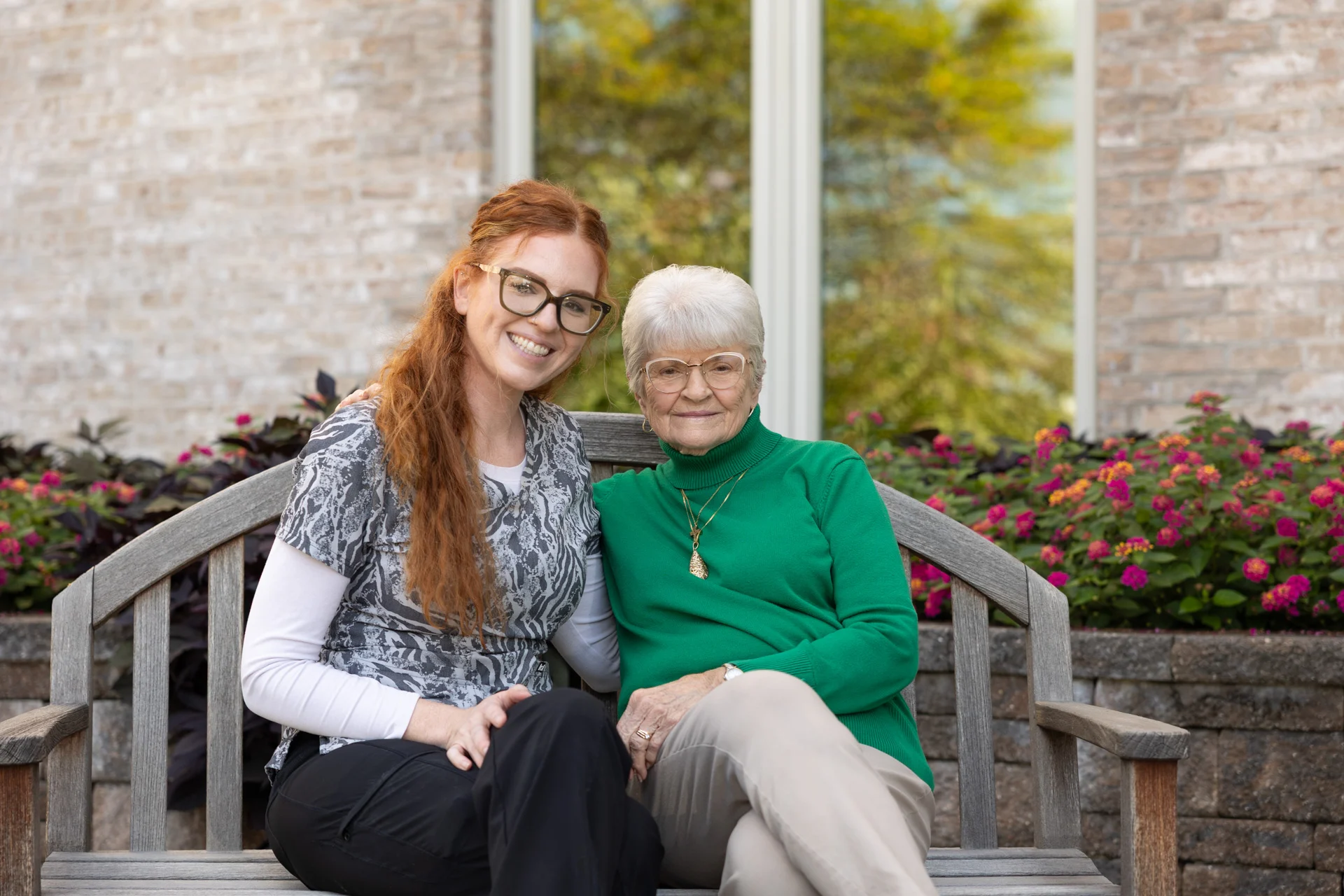1W8A0938 Smiling senior woman and nurse sitting on bench outdoors together.
